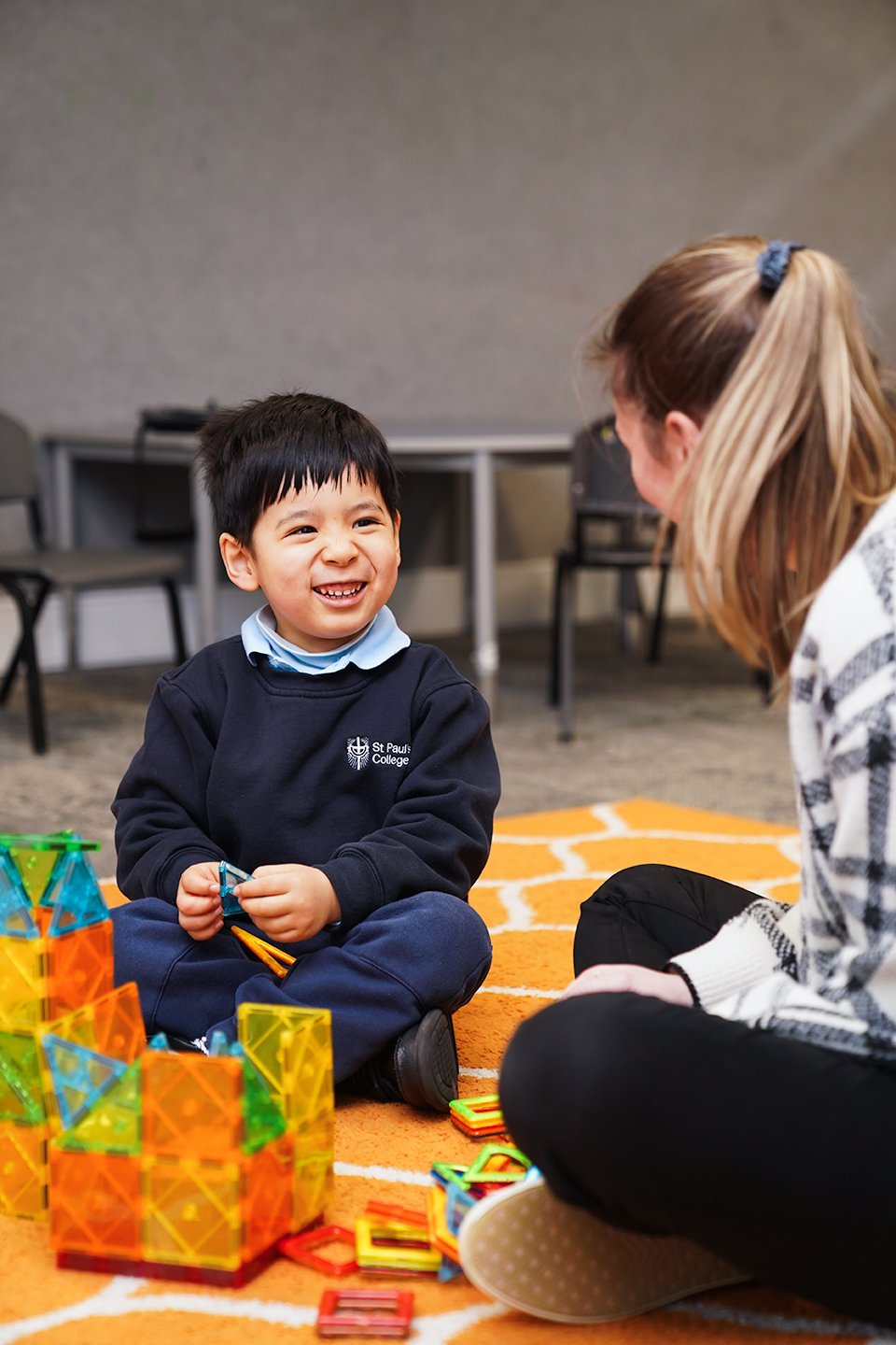 Boy sitting with his legs crossed on an orange carpet with building blocks, looking at a female teacher.