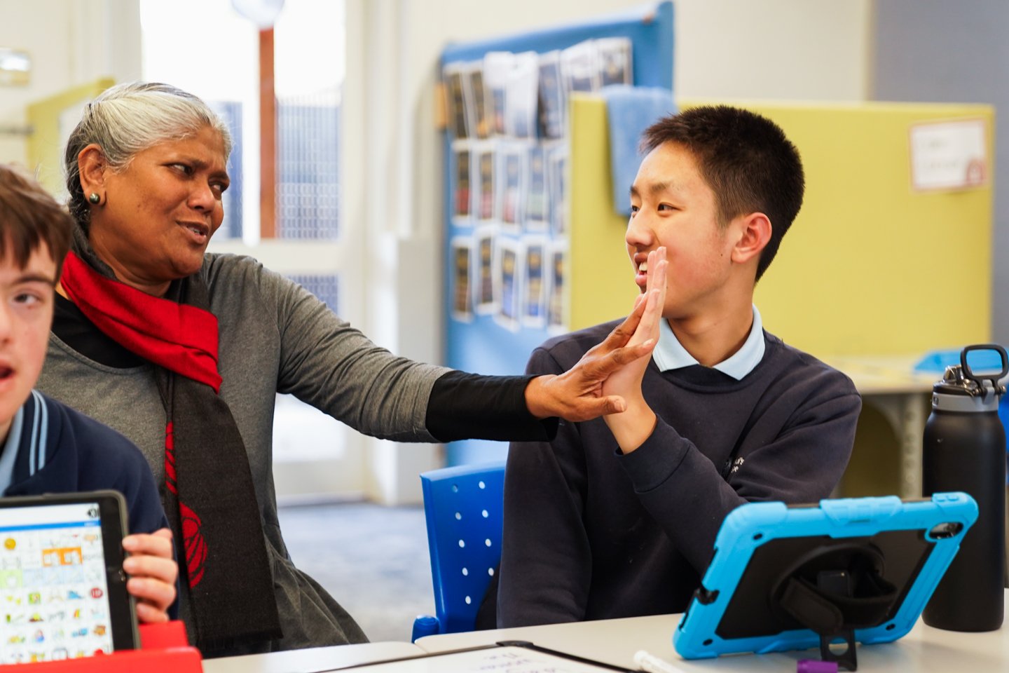 A woman on the right is giving a high-five to a male student with black hair wearing a navy uniform on a blue iPad device.