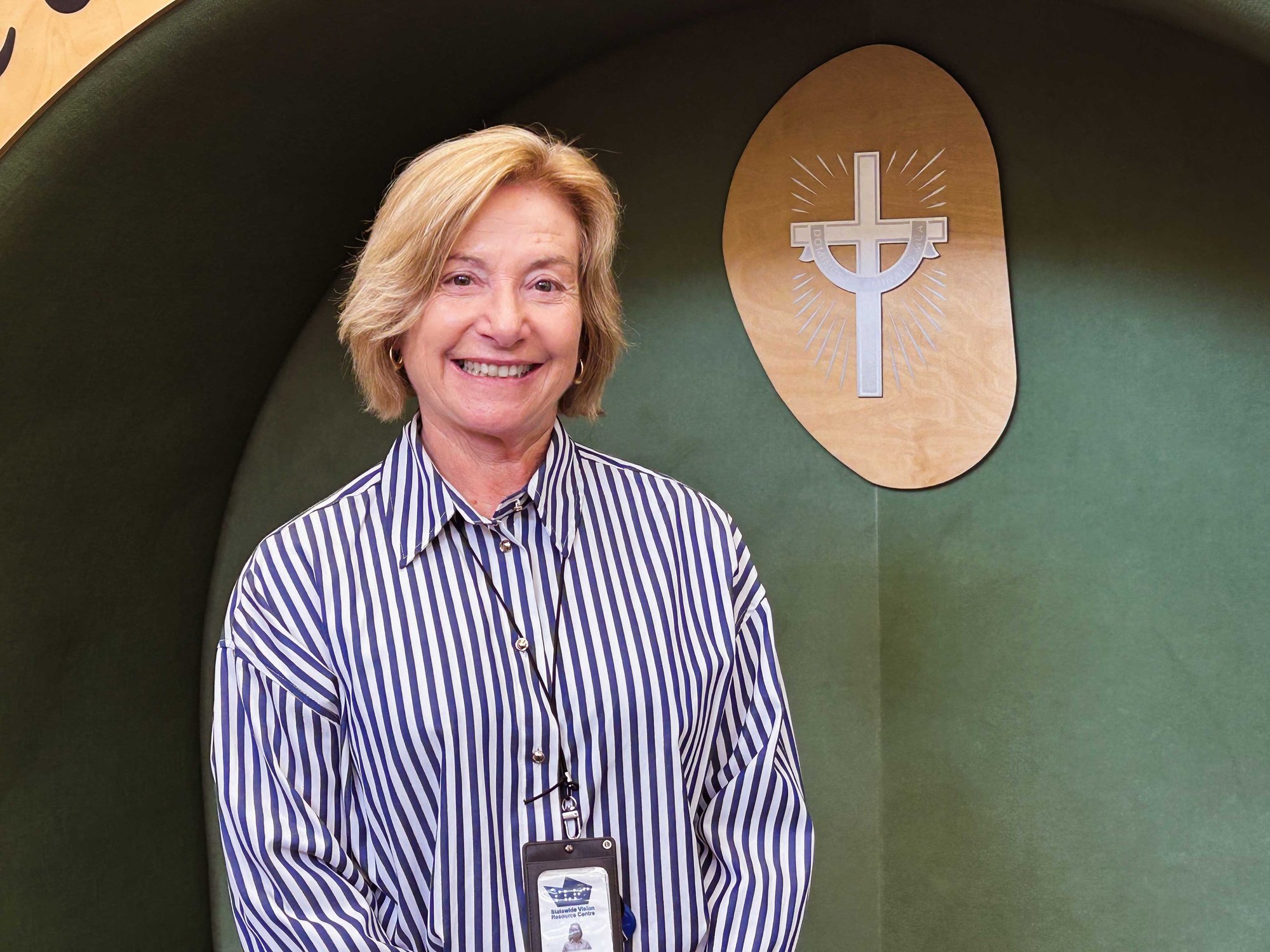 A blonde woman smiling at the camera, wearing a white-and-blue striped shirt, in front of a green background with a timber cross on the right-hand side.